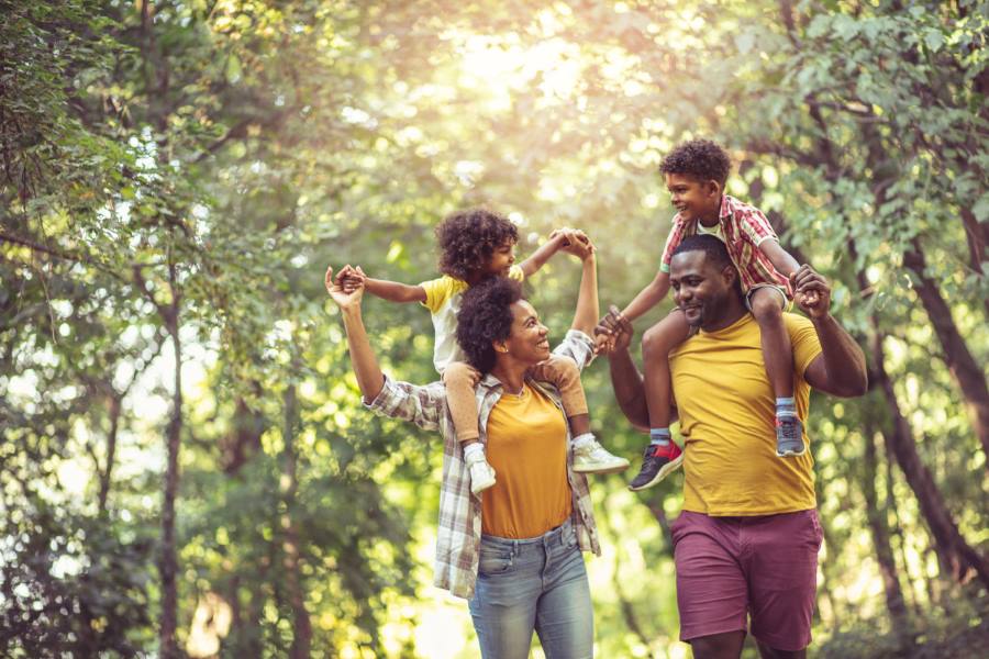 Two parents walk through a forest, each with a child sitting on their shoulders. The parents look at each other and smile.