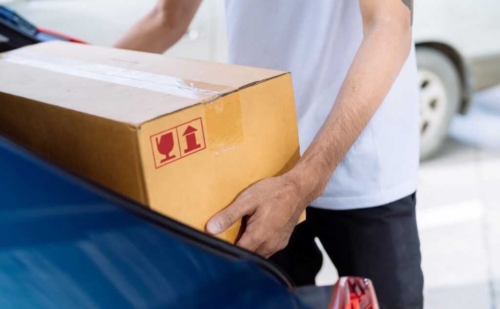 A man placing a taped cardboard box into the trunk of his car, preparing for transport to his storage unit.