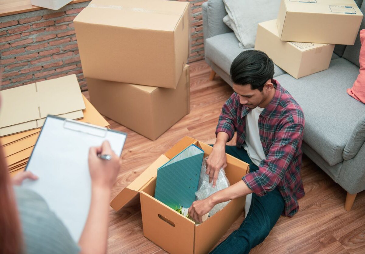 A man packing belongings in a cardboard box while a woman checks off items, surrounded by stacked moving boxes.