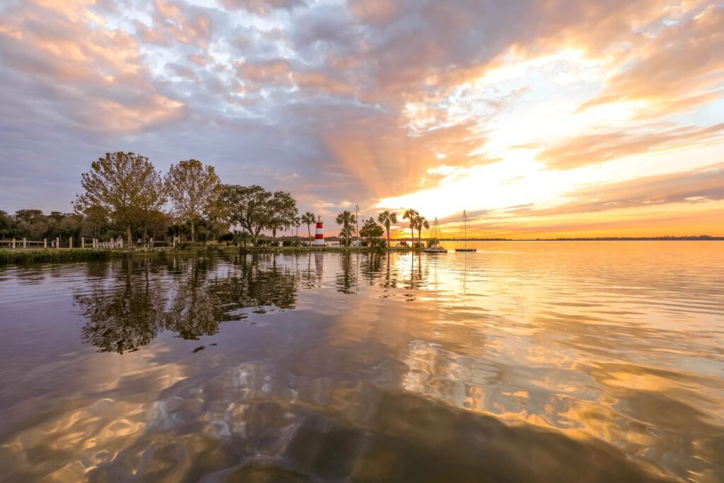 Scenic view of a peaceful Mount Dora, FL, waterfront at sunset, with calm water, trees, and vibrant rays of sunlight. 