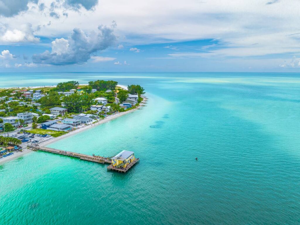 An aerial view of Anna Maria Island, FL, showcasing turquoise waters, a wooden pier, and charming homes. 