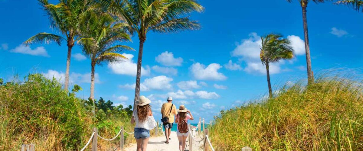 A group walking along a sandy path lined with palm trees toward South Beach, Miami, under a bright blue sky.