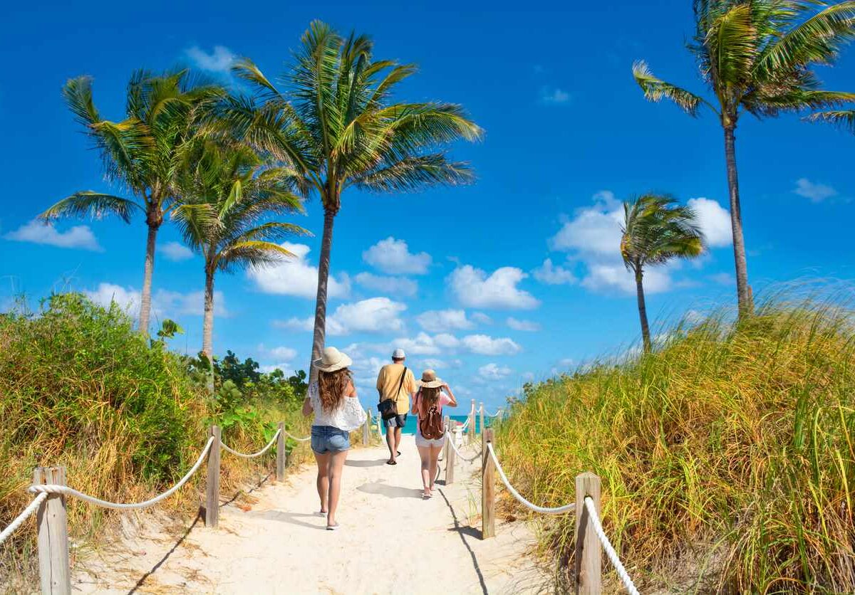 A group walking along a sandy path lined with palm trees toward South Beach, Miami, under a bright blue sky.