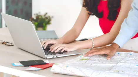 Two people planning travel using a laptop and a map, with passports and notebooks on the table.