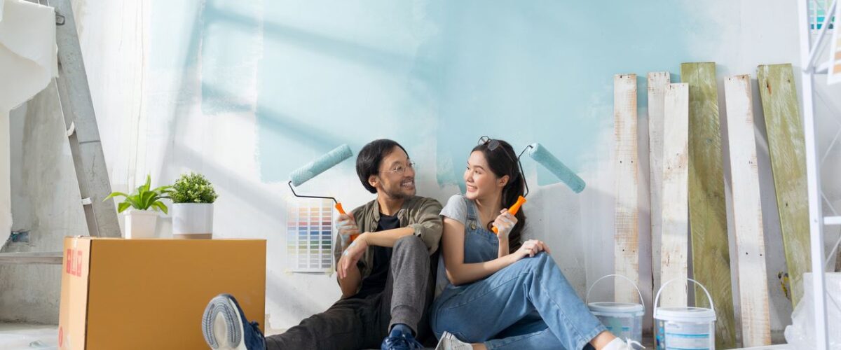 A happy couple sits on the floor, painting a light blue wall surrounded by paint supplies and wooden planks.