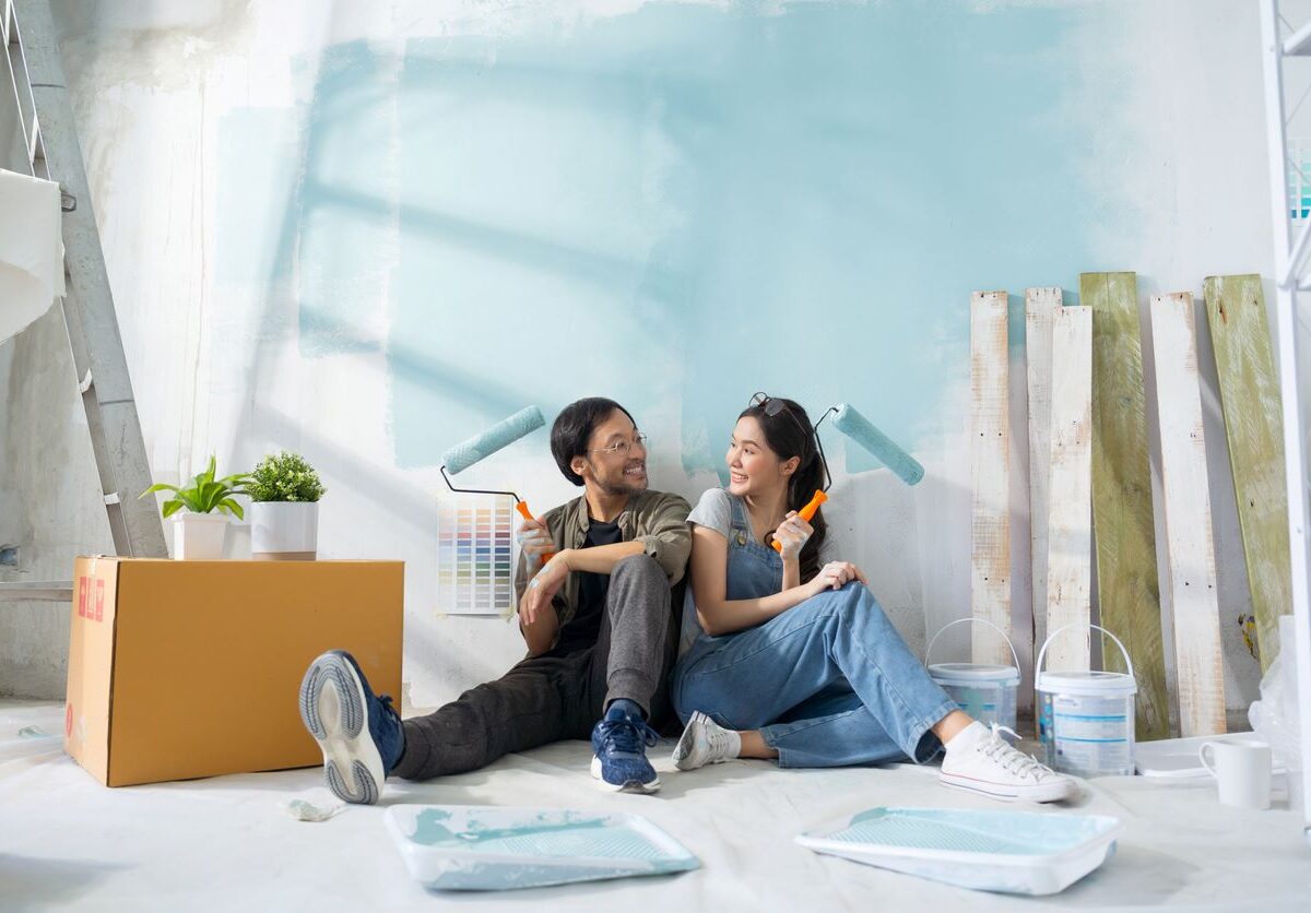 A happy couple sits on the floor, painting a light blue wall surrounded by paint supplies and wooden planks.