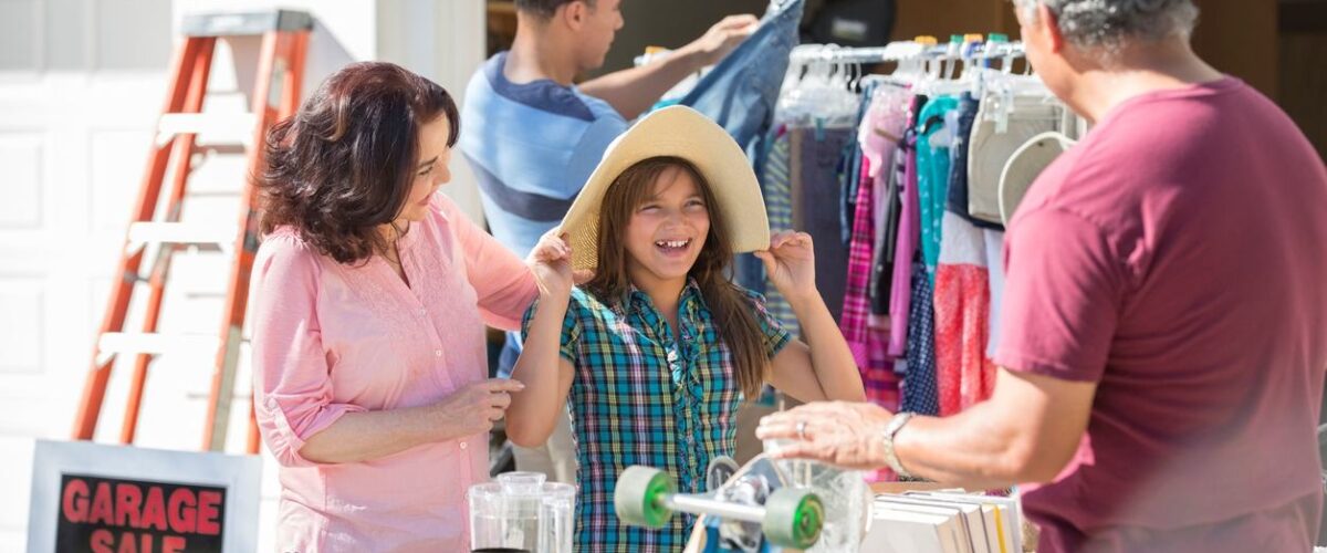 A family shops at a garage sale, with a young girl trying on a sun hat and tables full of items for sale.