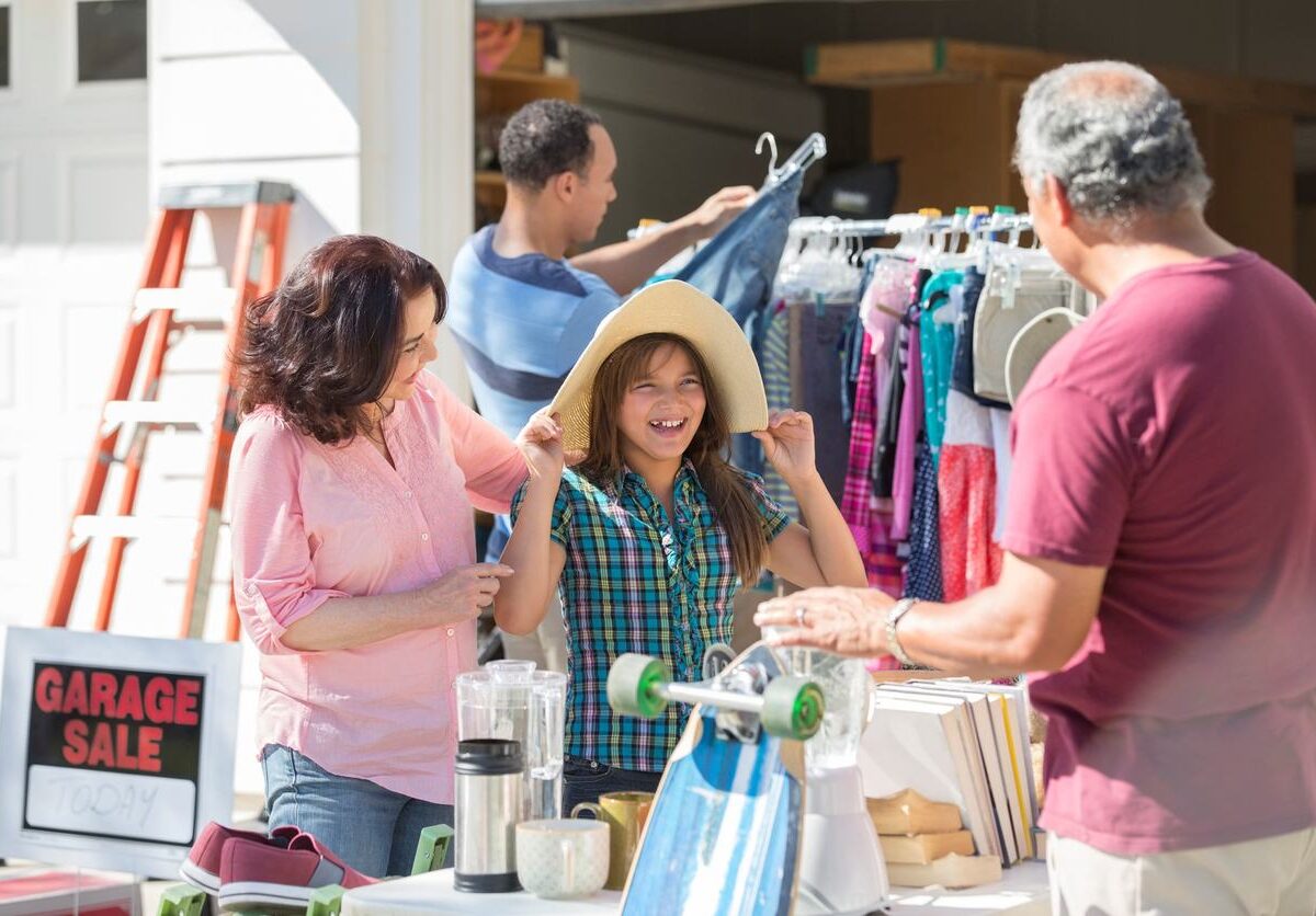 A family shops at a garage sale, with a young girl trying on a sun hat and tables full of items for sale.