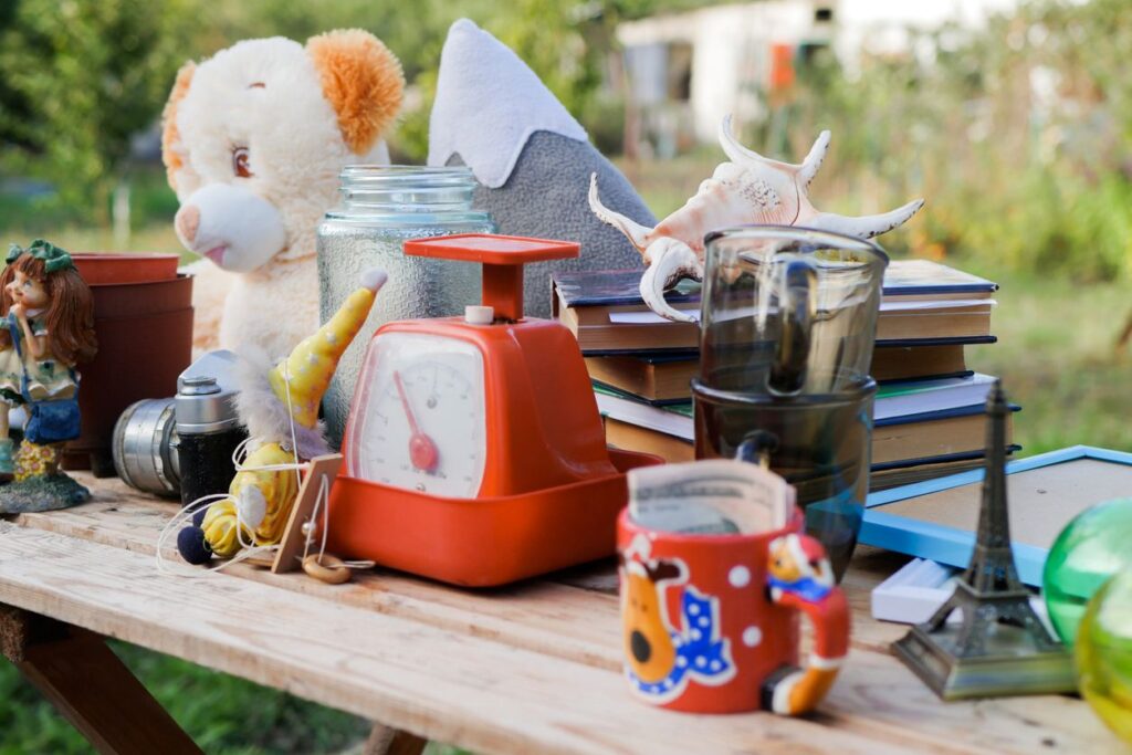 A table at a yard sale filled with assorted items, including toys, books, mugs, a scale, and knickknacks.