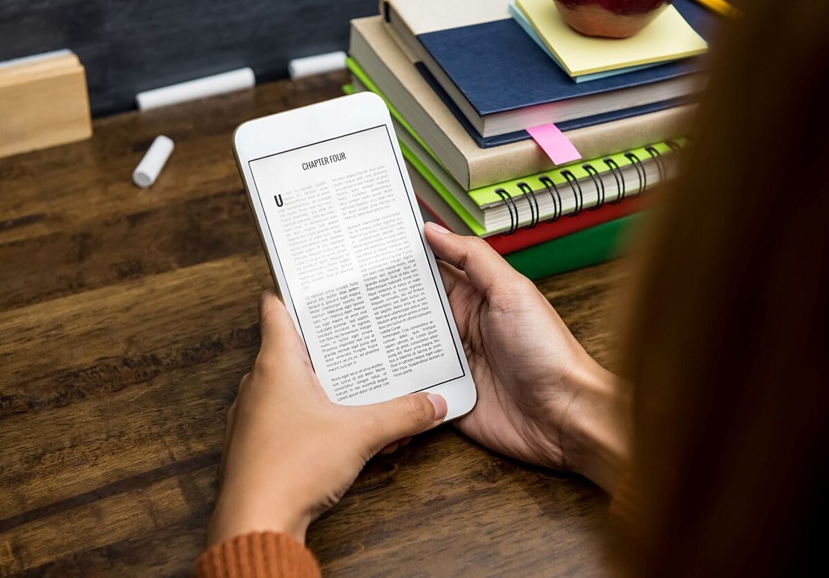 A woman reading an e-book on a smartphone at a wooden desk with stacked books, notebooks, and an apple.