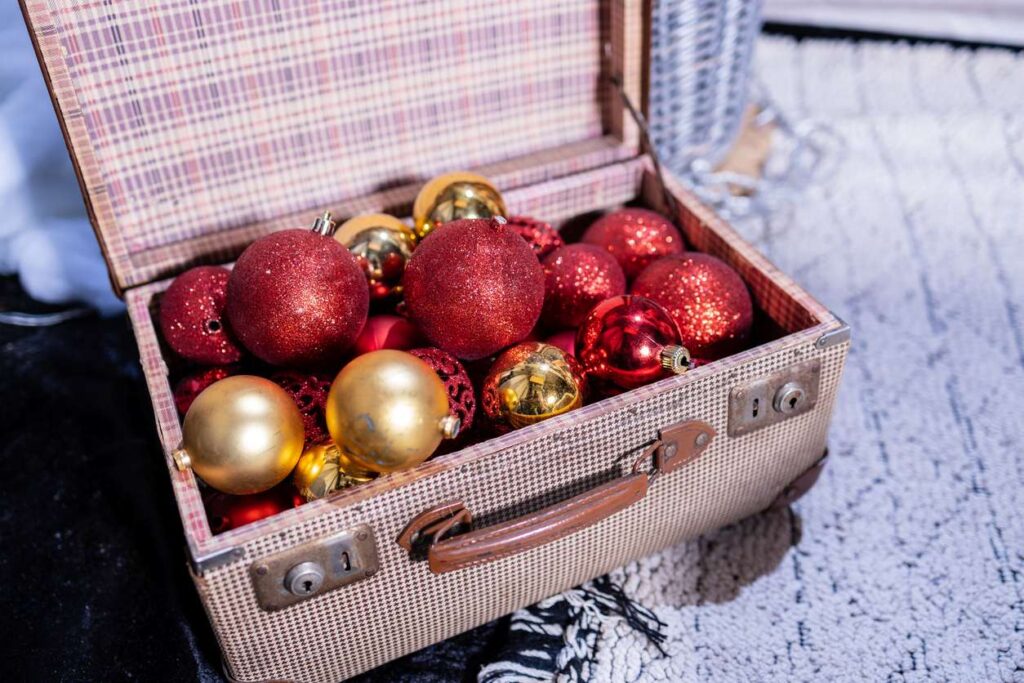 An open vintage suitcase filled with red and gold holiday ornaments, sitting on a soft rug and ready to be packed away.
