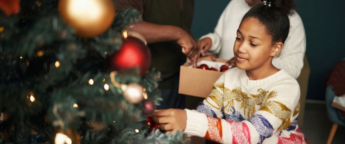 Family packing holiday decorations into labeled storage boxes beside a Christmas tree in a living room.